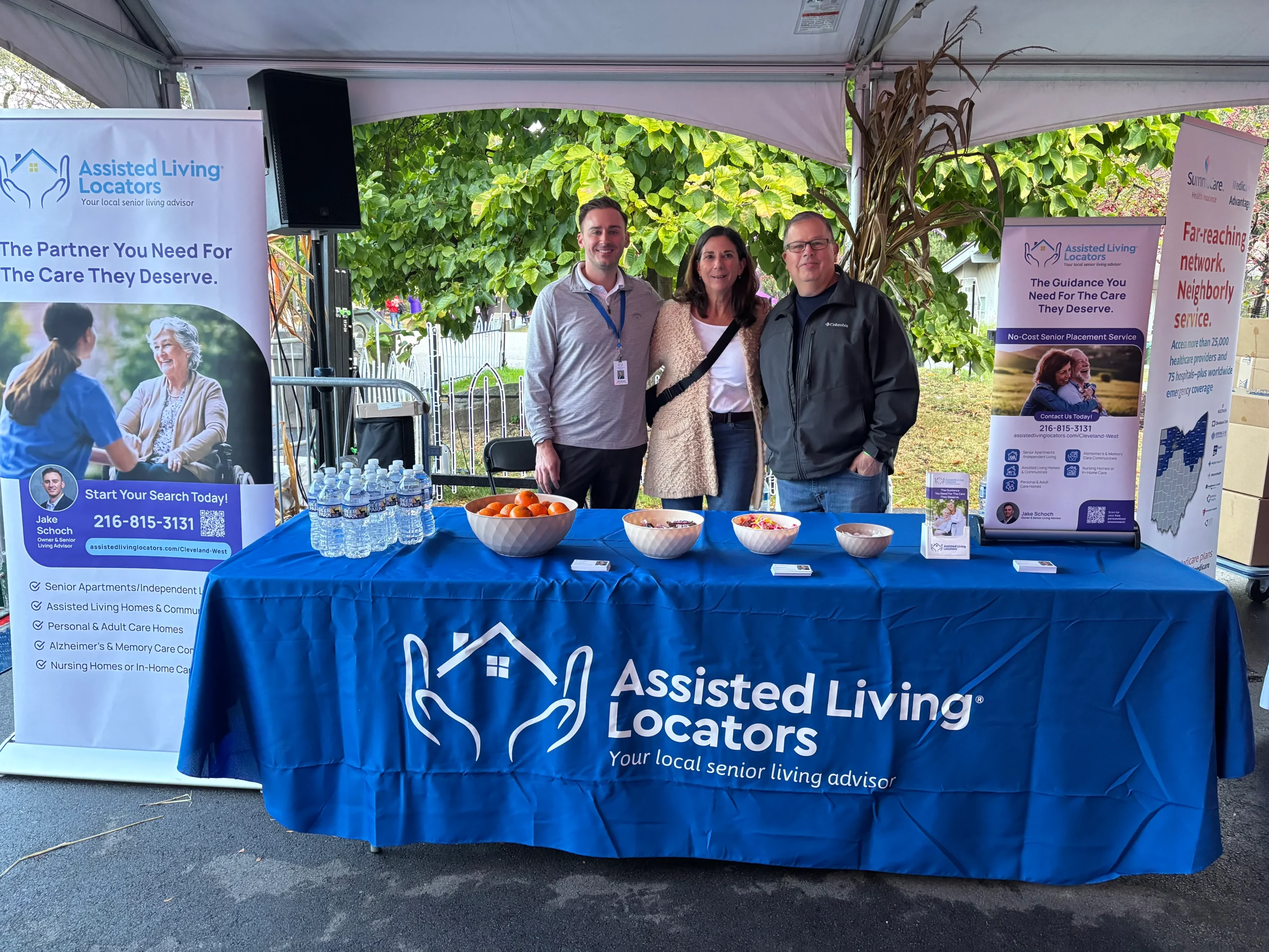 Three Assisted Living Locators team members standing behind a branded booth at a community outreach event with brochures, snacks, and promotional banners displayed.