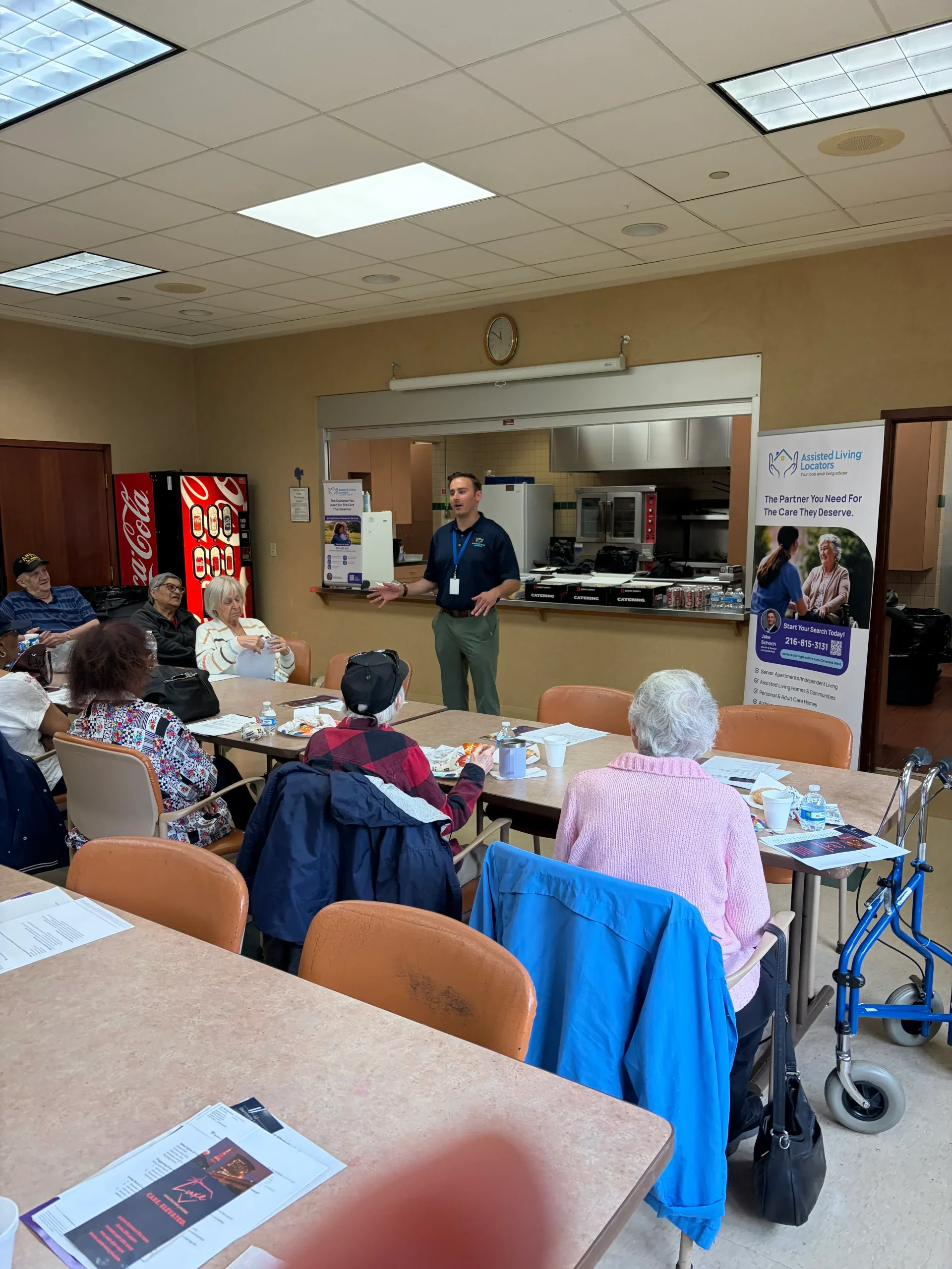 Senior residents attending an informational session in a dining area with an Assisted Living Locators representative presenting at the front of the room.