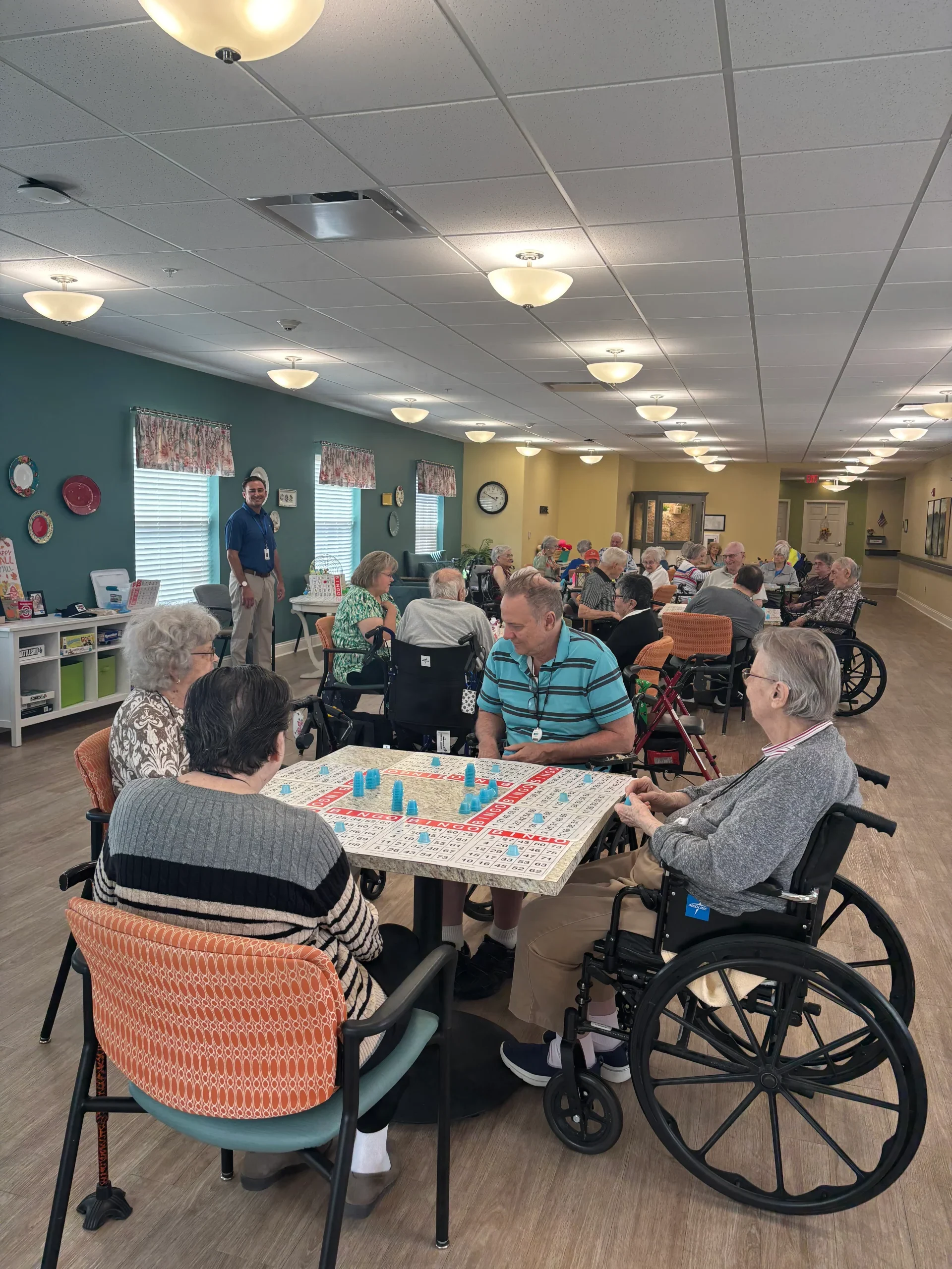 Residents gathered in a community room playing bingo at an assisted living facility while a staff member speaks to the group.
